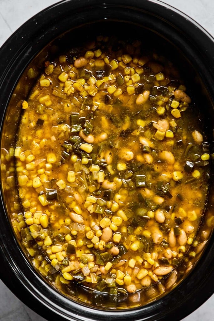 Slow cooker filled with a cooked mixture of white beans, corn, bell peppers, and onions in a broth.