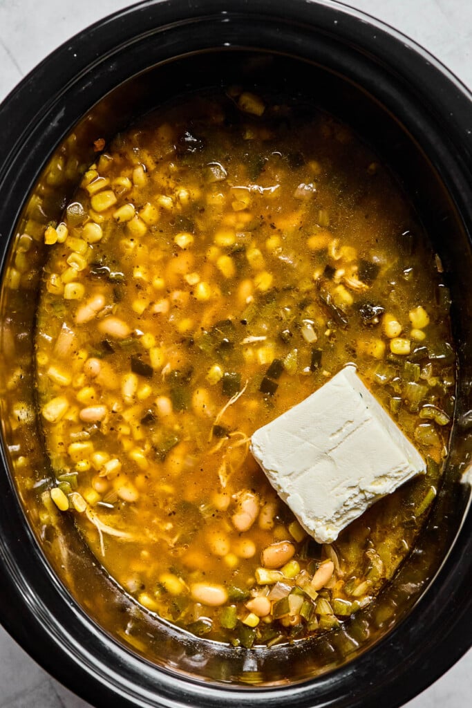 A slow cooker filled with soup with a brick of cream cheese sitting on top of it before it is mixed it.