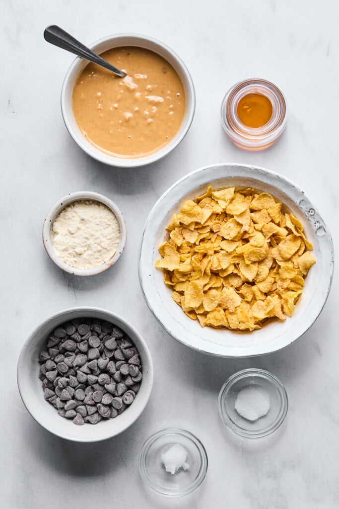 Small jar of honey, a bowl of cornflakes, a small bowl of salt, a small bowl of coconut oil, a bowl of chocolate chips, a small bowl of coconut flour, and a bowl of crunchy peanut butter with a spoon in on a marble counter. 