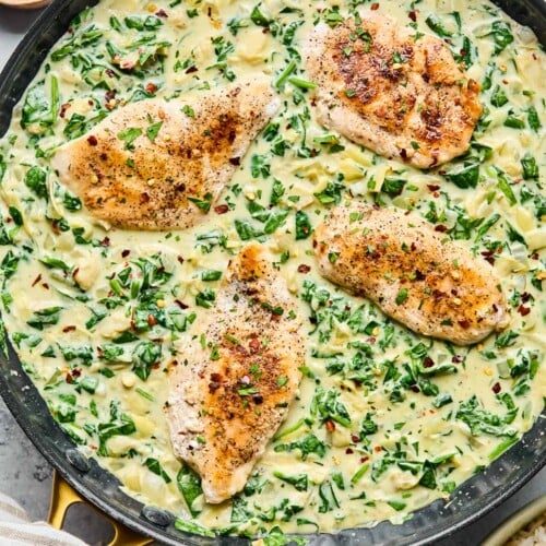Spinach artichoke chicken skillet garnished with red pepper flakes. Next to the pan is a bowl of rice, a striped kitchen towel, a small bowl of red pepper flakes, and a small bowl of parsley.