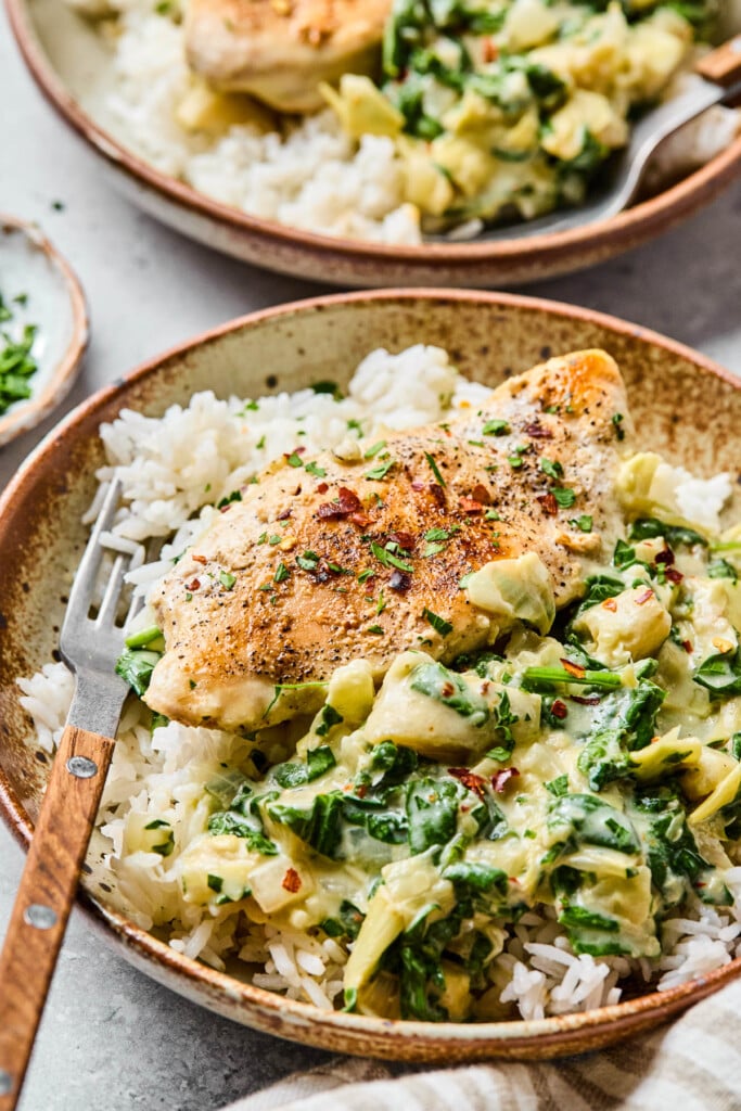 Spinach and artichoke chicken over a bed of white rice in a bowl. It is garnished with red pepper flakes and parsley and a fork is in the bowl. Around the bowl is a striped kitchen towel, a plate of chopped parsley, and another bowl of spinach and artichoke chicken.