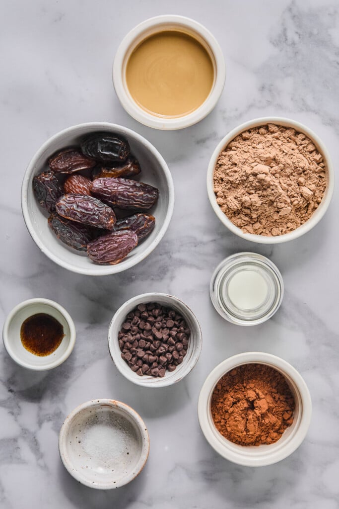 A bowl of tahini, a bowl of chocolate protein powder, a jar of milk, a bowl of cocoa powder, a bowl of flaky sea salt, a bowl of mini chocolate chips, a small bowl of vanilla bean paste, and a bowl of medjool dates on a marble counter.
