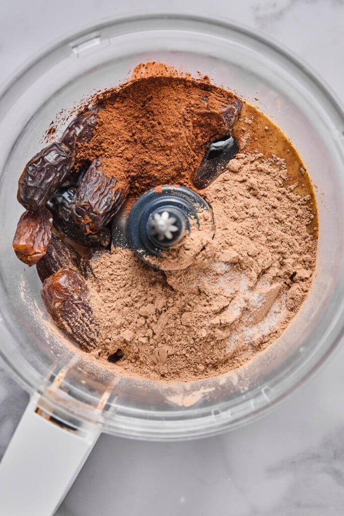 Food processor filled with medjool dates, tahini, cocoa powder, chocolate protein powder, and salt.