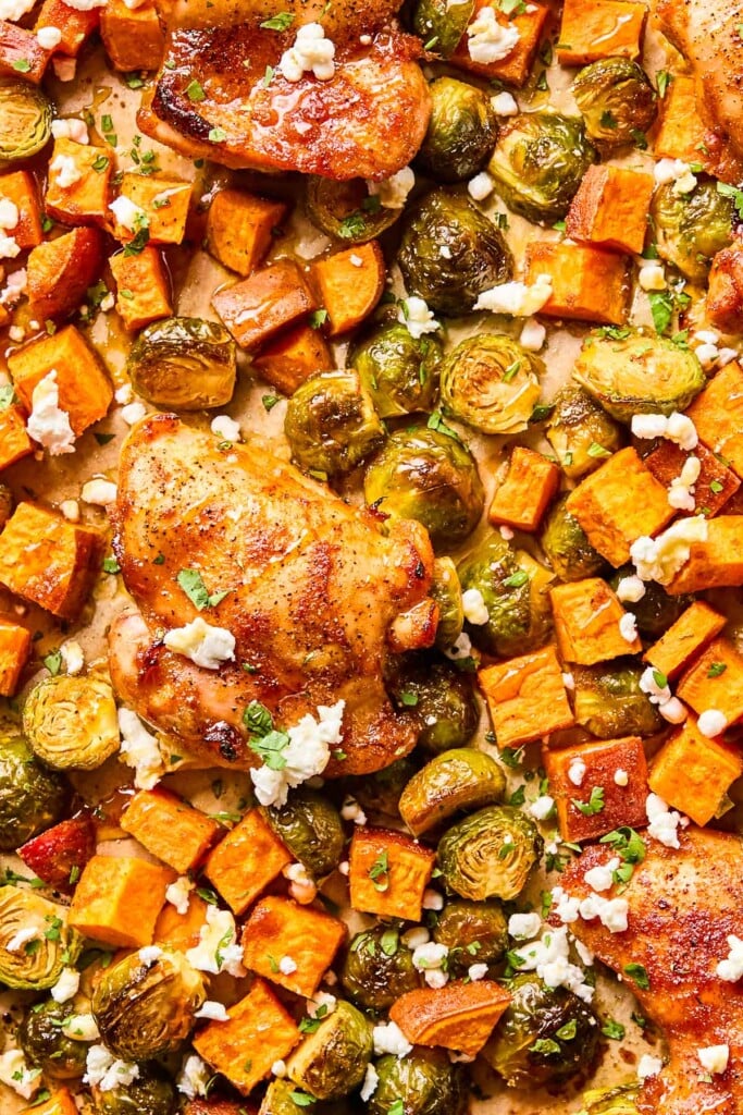 A pan of baked hot honey chicken with roasted sweet potatoes and brussels sprouts. It is garnished with goat cheese and chopped cilantro.