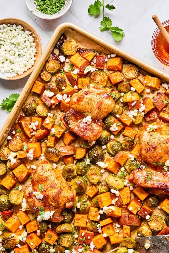 Hot honey chicken thighs with sweet potatoes, brussels sprouts, and goat cheese on a sheet pan. Everything is garnished with cilantro. Around the pan is a bowl of goat cheese, a bowl of chopped cilantro and a bowl of hot honey with a honey dipper in it.