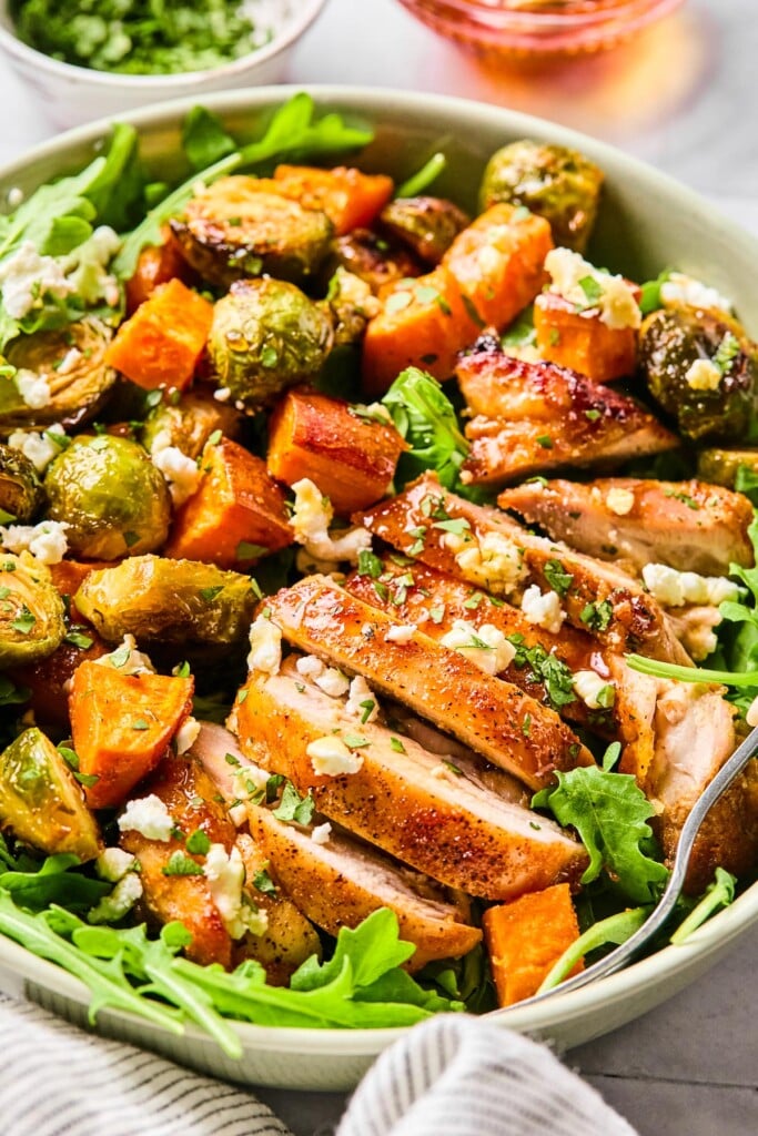 Hot honey chicken bowls with arugula, roasted sweet potatoes, roasted brussel sprouts, goat cheese, and cilantro. Around the bowl is striped kitchen towel, a bowl of chopped cilantro, and a bowl of hot honey.