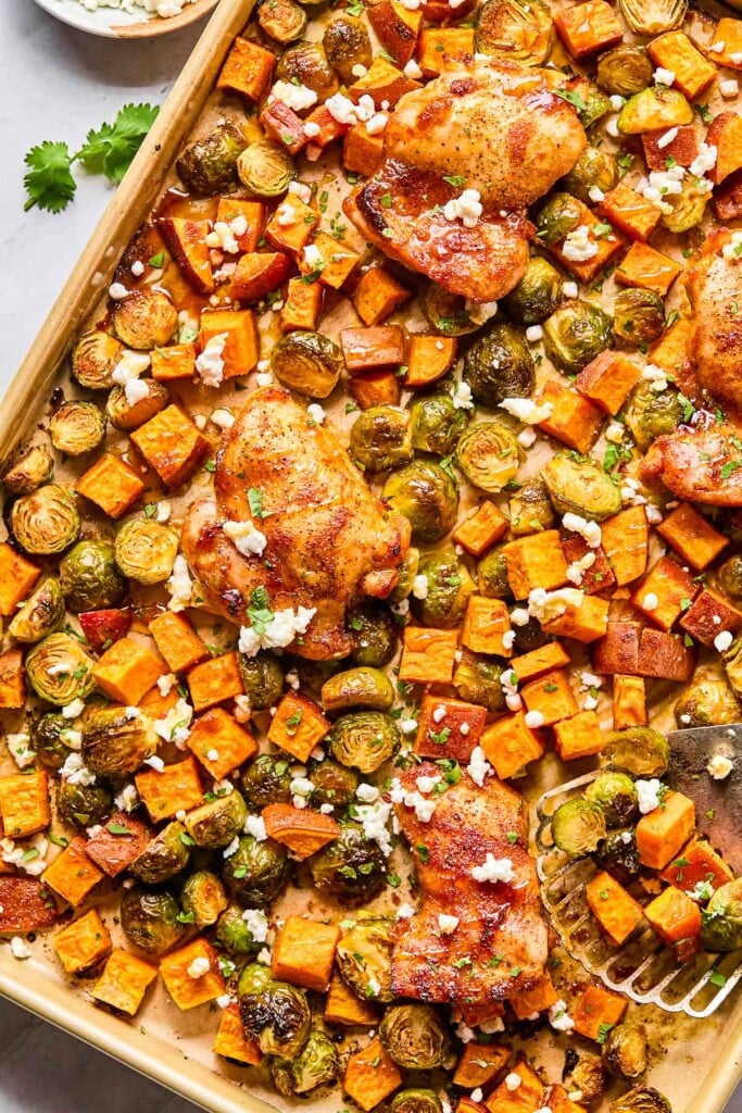 Sheet pan hot honey chicken with sweet potatoes and brussels sprouts topped with cilantro and goat cheese. A metal spatula is on the sheet pan with a few veggies on it.