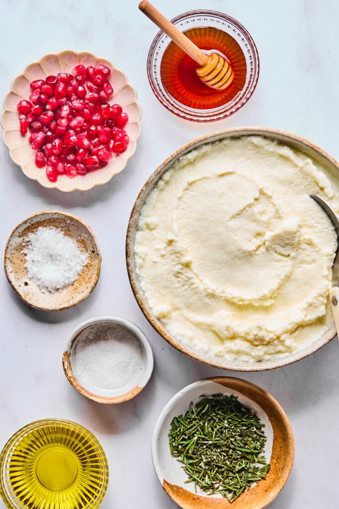 A small bowl with hot honey with a honey dipper in it, a bowl of ricotta cheese with a spoon in it, a bowl of fresh chopped rosemary, a small bowl with olive oil, a small bowl of sea salt, a small dish of flaky sea salt, and a dish of pomegranate seeds on a white counter.