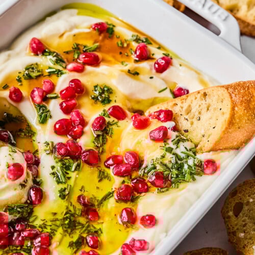 Whipped ricotta dip in a white serving dish topped with olive oil, fresh rosemary, pomegranate seeds, and hot honey. A piece of crostini is dipped into the ricotta and around the pan is more crostini.