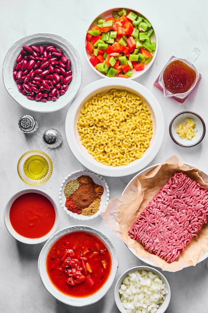 A bowl of diced red and green bell peppers, a cup on bone broth, a bowl of uncooked macaroni, a small dish of minced garlic, a plate of a pound of raw ground beef, a bowl of diced onions, a bowl of fire-roasted tomatoes in sauce, a bowl of tomato sauce, a plate of different spices, a small dish of olive oil, salt and pepper shakers, and a bowl of red kidney beans on a grey counter.