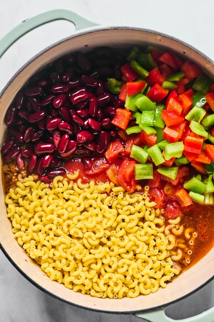 Overhead shot of a large Dutch oven filled with uncooked macaroni pasta, seasoned ground beef, diced green and red bell peppers, kidney beans, tomato sauce, and broth before it is mixed together.