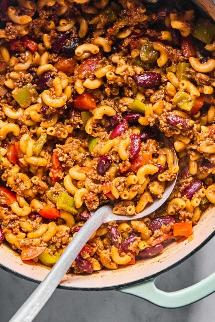 Large Dutch oven filled with a mixture of cooked meat, veggies, beans, and pasta in sauce. A metal spoon is in the pot.
