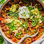 Overhead shot of chili pasta in a large Dutch oven. It is topped with sour cream, shredded cheese, sliced avocado, sliced jalape&ntilde;os, sliced green onions, and cilantro. A metal spoon is also in the pot. Around the pot is a striped kitchen towel, a bowl of sliced green onions, a bowl of sliced jalape&ntilde;os, a bowl of shredded cheese, and a bowl of chopped cilantro.