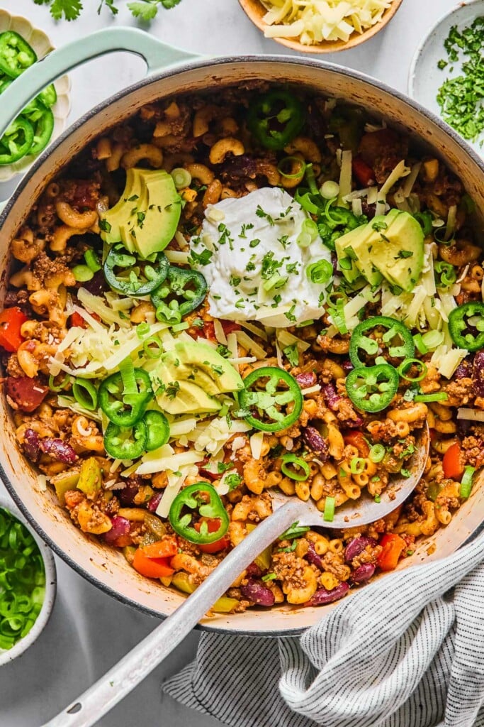Overhead shot of chili pasta in a large Dutch oven. It is topped with sour cream, shredded cheese, sliced avocado, sliced jalapeños, sliced green onions, and cilantro. A metal spoon is also in the pot. Around the pot is a striped kitchen towel, a bowl of sliced green onions, a bowl of sliced jalapeños, a bowl of shredded cheese, and a bowl of chopped cilantro.