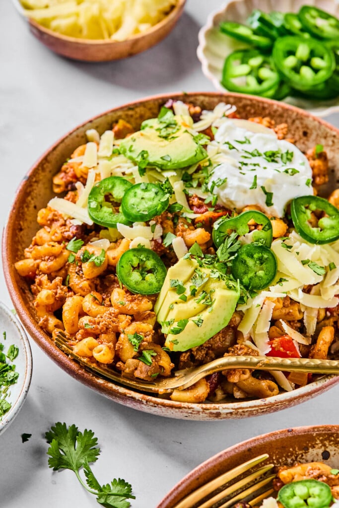 Chili with pasta topped with sour cream, shredded cheese, sliced avocado, sliced jalape&ntilde;os, and cilantro in a bowl. A gold fork is also in the bowl. Around the bowl is another bowl of chili, some cilantro leaves, a bowl of shredded cheese, and a bowl of sliced jalape&ntilde;os.