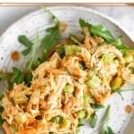 Healthy chicken salad with celery, carrots, and buffalo sauce on top of some arugula on a piece of bread on a plate. It is drizzled with hot sauce. Around the plate is a plate of carrots and celery and a bowl of more chicken salad.