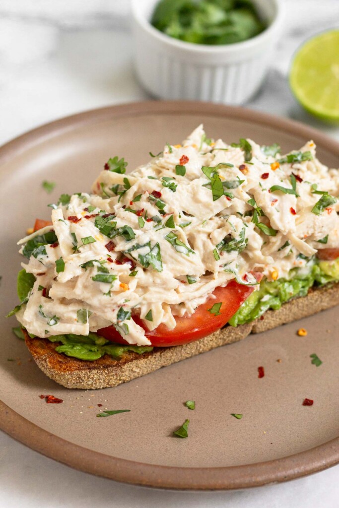 A piece of toast with mash avocado, sliced tomato, and cilantro lime chicken salad on top of it on a brown plate. Behind the plate is a small dish of cilantro and half a lime.