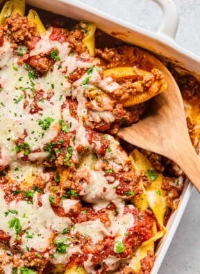 Overhead shot of a pan filled with stuffed shells with meat sauce topped with melted cheese, parsley, and parmesan cheese. There is a wooden spoon in the pan with a stuffed shell on it.
