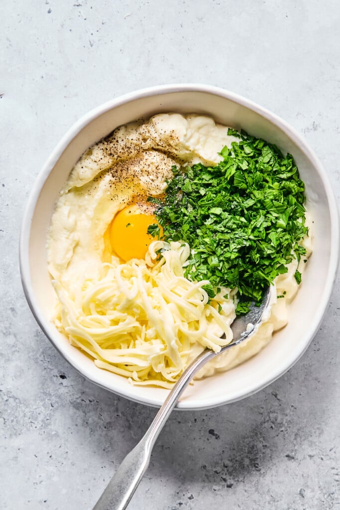 Overhead shot of a bowl with ricotta cheese, shredded mozzarella cheese, an egg, fresh chopped parsley, and black pepper before it is mixed together. There is a spoon in the spoon in the bowl as well.