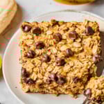 A piece of banana baked oatmeal with chocolate chips on a white plate. A little piece is taken off the corner and sitting on a fork on the plate. Around the plate are some chocolate chips and some bananas.