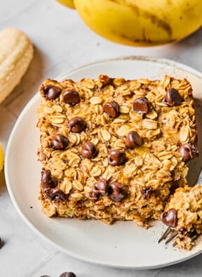 A piece of banana baked oatmeal with chocolate chips on a white plate. A little piece is taken off the corner and sitting on a fork on the plate. Around the plate are some chocolate chips and some bananas.