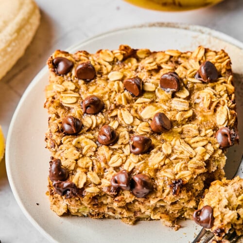 A piece of banana baked oatmeal with chocolate chips on a white plate. A little piece is taken off the corner and sitting on a fork on the plate. Around the plate are some chocolate chips and some bananas.