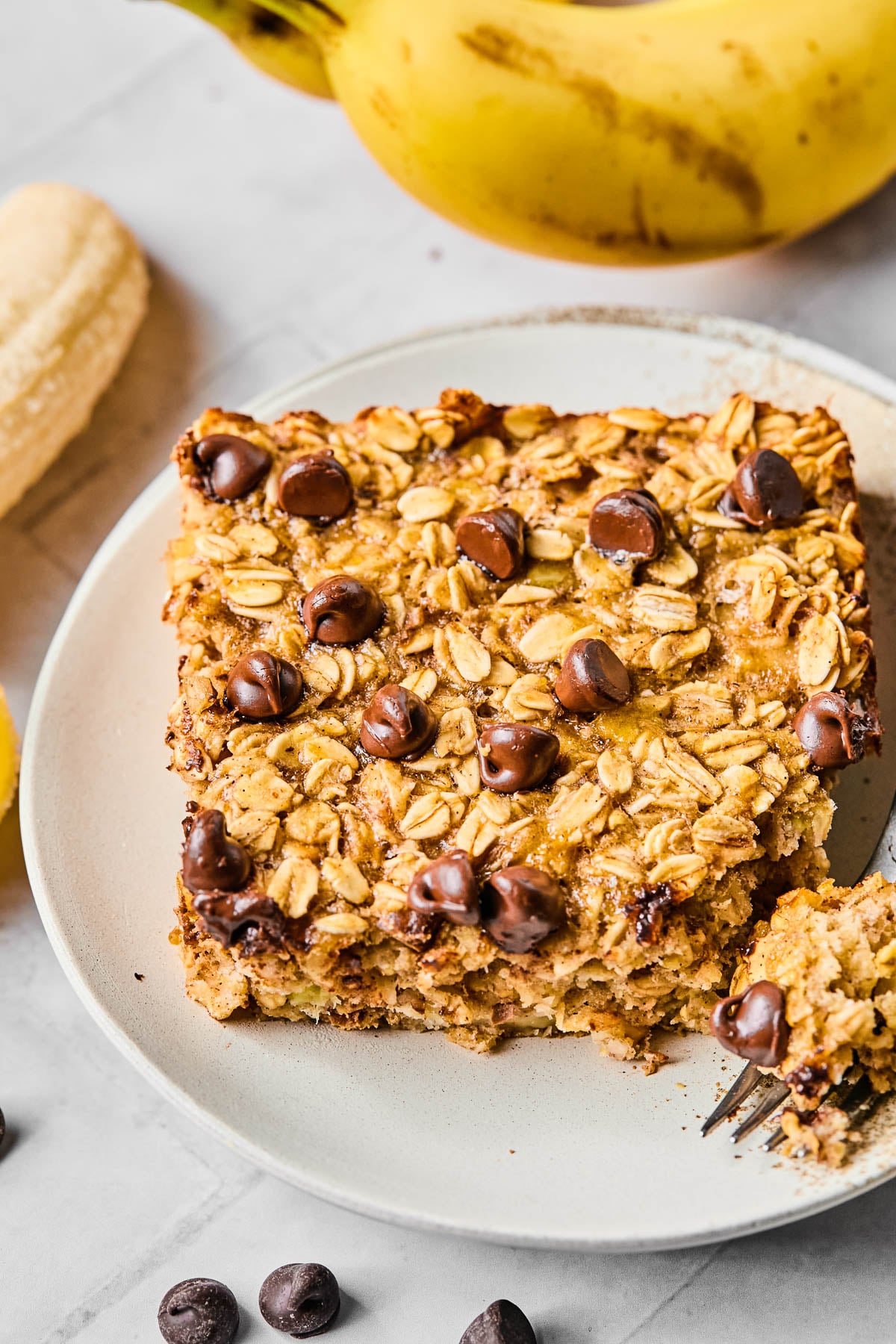 A piece of banana baked oatmeal with chocolate chips on a white plate. A little piece is taken off the corner and sitting on a fork on the plate. Around the plate are some chocolate chips and some bananas.