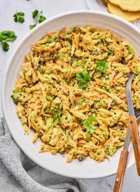 A large bowl of curry chicken salad with raisins, cashews, and veggies, garnished with cilantro. There is a spoon and a fork in the bowl as well. Around the bowl is a striped kitchen towel, some cilantro leaves, and some slices of bread.