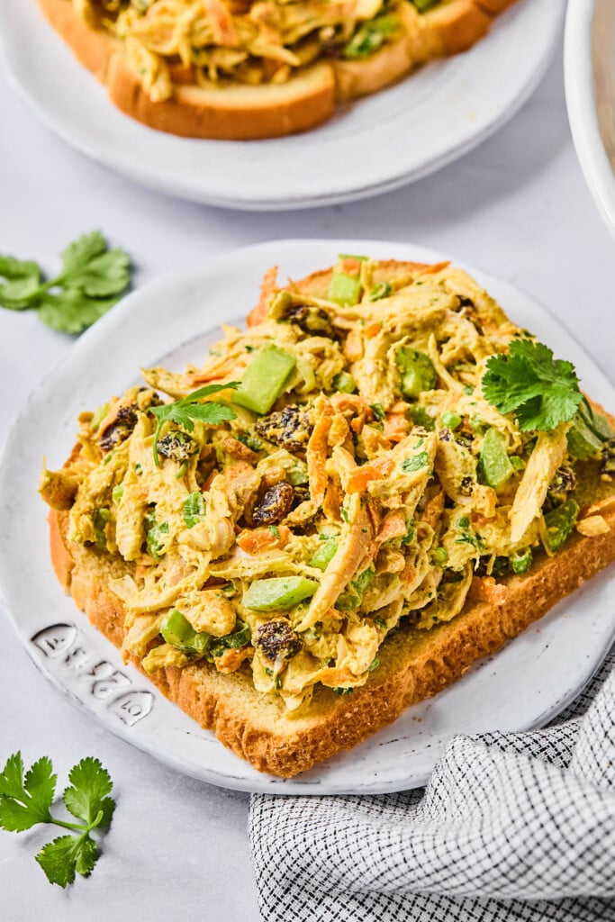Curry chicken salad with greek yogurt, cashews, and raisins on top of a piece of toast on a plate. It is garnished with fresh cilantro. Around the plate is a checkered kitchen towel, some fresh cilantro, and another plate of curry chicken salad on toast.
