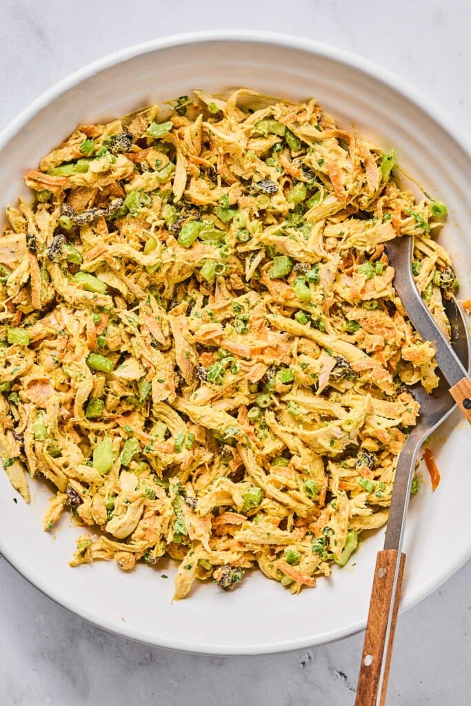 Healthy curry chicken salad with greek yogurt, raisins, cashews, veggies, green onions, and cilantro in a white bowl with a fork and spoon in it.