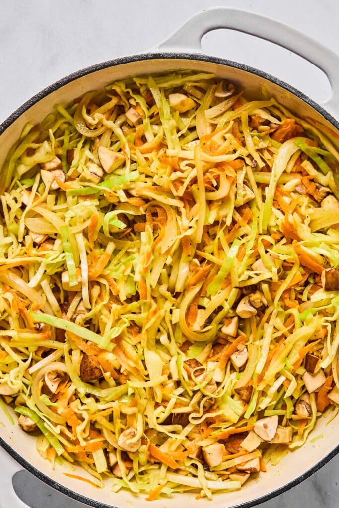 Overhead shot of a large white enameled pan filled with saut&eacute;ed sliced cabbage, shredded carrots, and diced mushrooms.