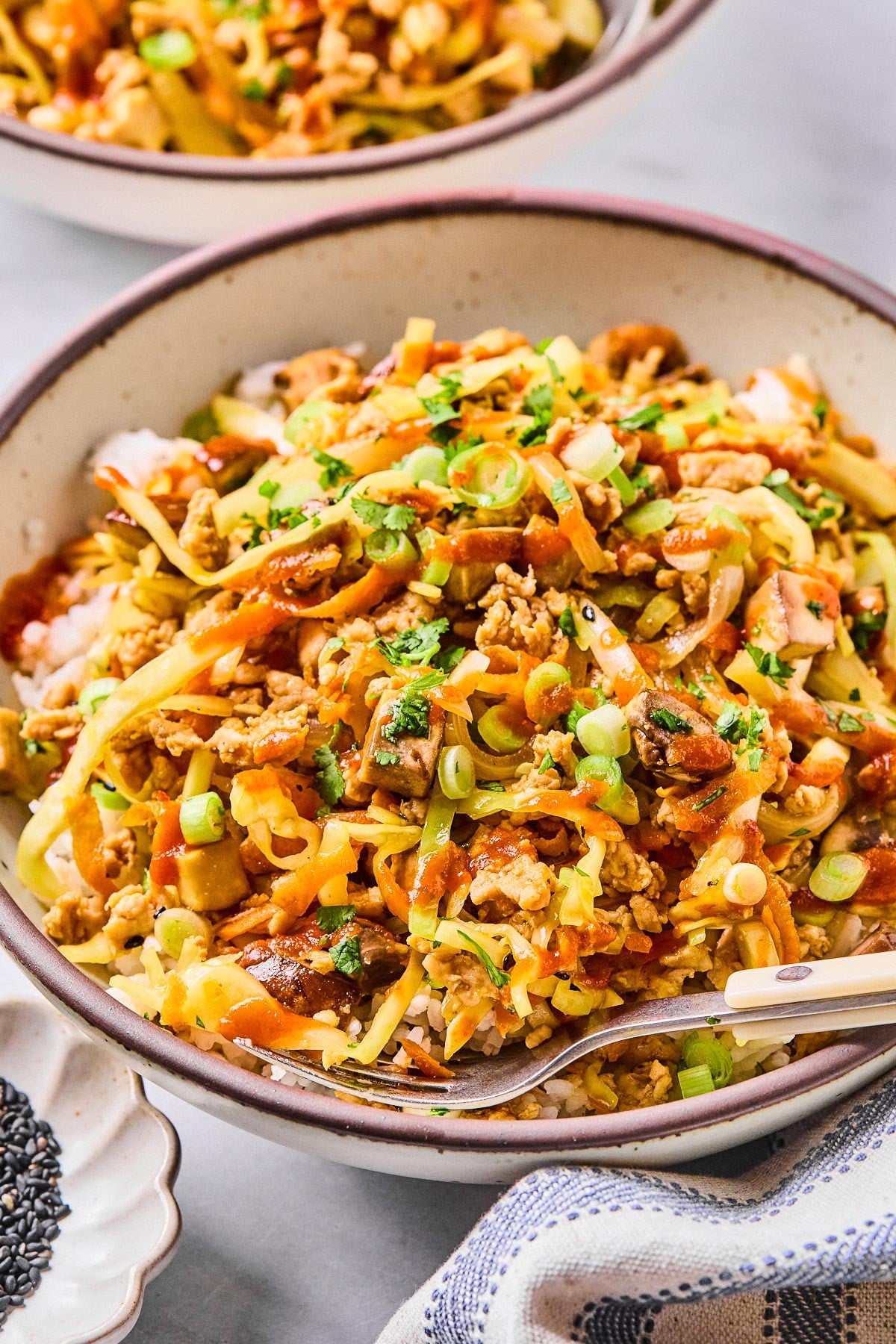Egg roll bowl with ground chicken, sliced cabbage, shredded carrots, and mushrooms over white rice. It is in a bowl and garnished with sriracha, green onions, cilantro, and sesame seeds. A fork is also in the bowl. Around the bowl is a kitchen towel, a bowl of black sesame seeds, and another egg roll bowl.