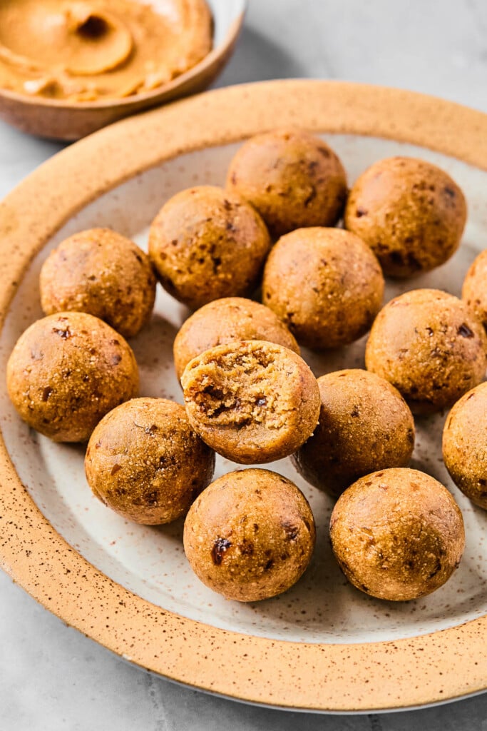 A plate of protein peanut butter balls with one of the balls stacked on top of the others, with a bite taken out of it. Behind the plate is a bowl of peanut butter.