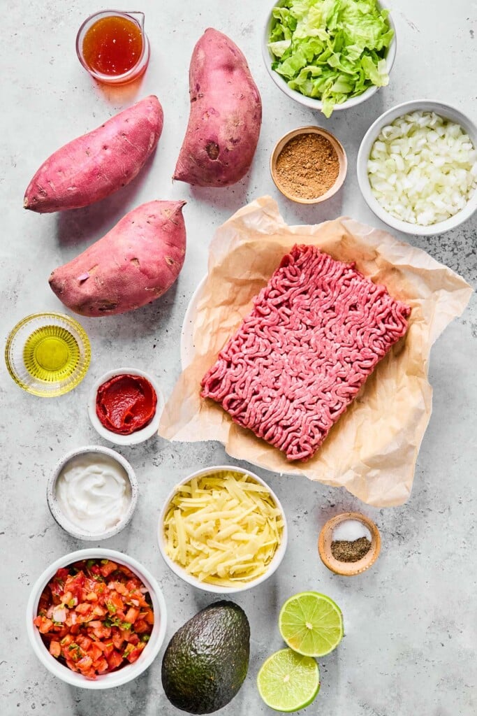 A bowl of shredded lettuce, a bowl of diced onions, a small bowl of taco seasoning, a plate with a pound of raw ground beef on parchment paper, a small bowl of salt and pepper, a bowl of shredded cheese, a lime that has been cut in half, a whole avocado, a bowl of pico de gallo, a small bowl of greek yogurt, a small bowl of tomato paste, a small cup of olive oil, 3 uncooked sweet potatoes, and a small pitcher of broth on a grey concrete counter.