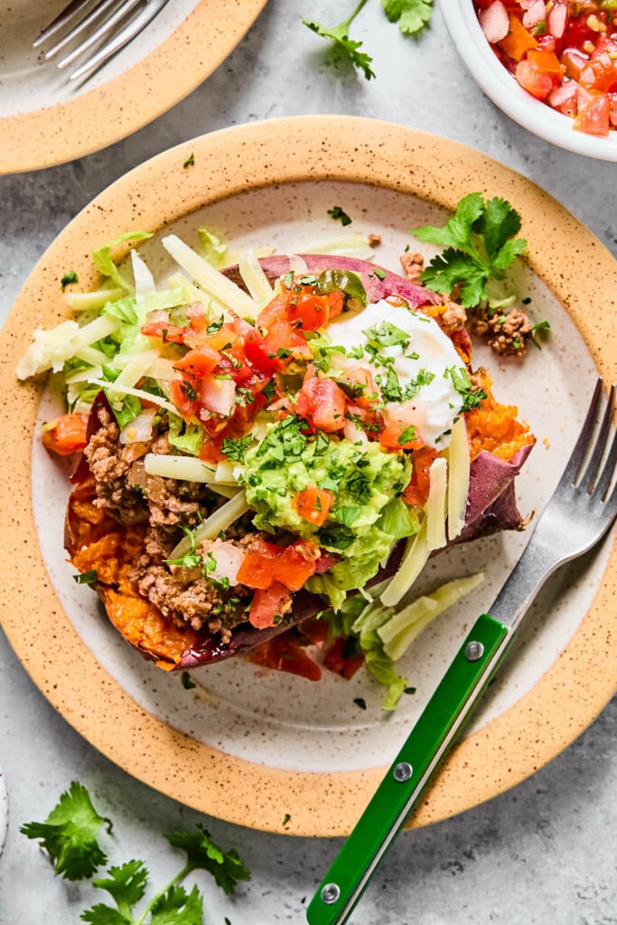 A taco stuffed sweet potato with taco meat, shredded cheese, shredded lettuce, guacamole, pico de gallo, greek yogurt, and cilantro on a plate. There is a fork on the plate as well. Around the plate are some cilantro leaves, a bowl of pico de gallo, and another plate.