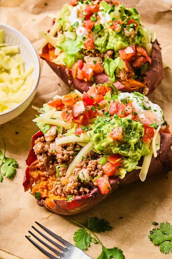 Two taco stuffed sweet potatoes piece of parchment paper. They are stuffed with taco meat, shredded cheese, shredded lettuce, pico de gallo, guacamole, greek yogurt, and cilantro. Around them is a fork, some cilantro leaves, and a bowl of shredded cheese.