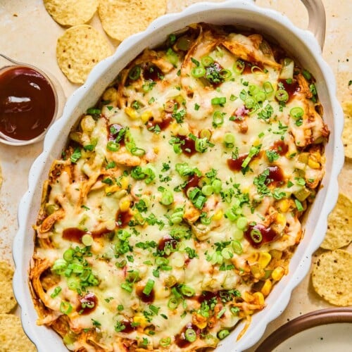 Au gratin dish filled with BBQ chicken dip topped with melted cheese and garnished with sliced green onions, cilantro, and BBQ sauce. Around the dish are some tortilla chips, an empty plate, a cup of bbq sauce, and a plate of cilantro.