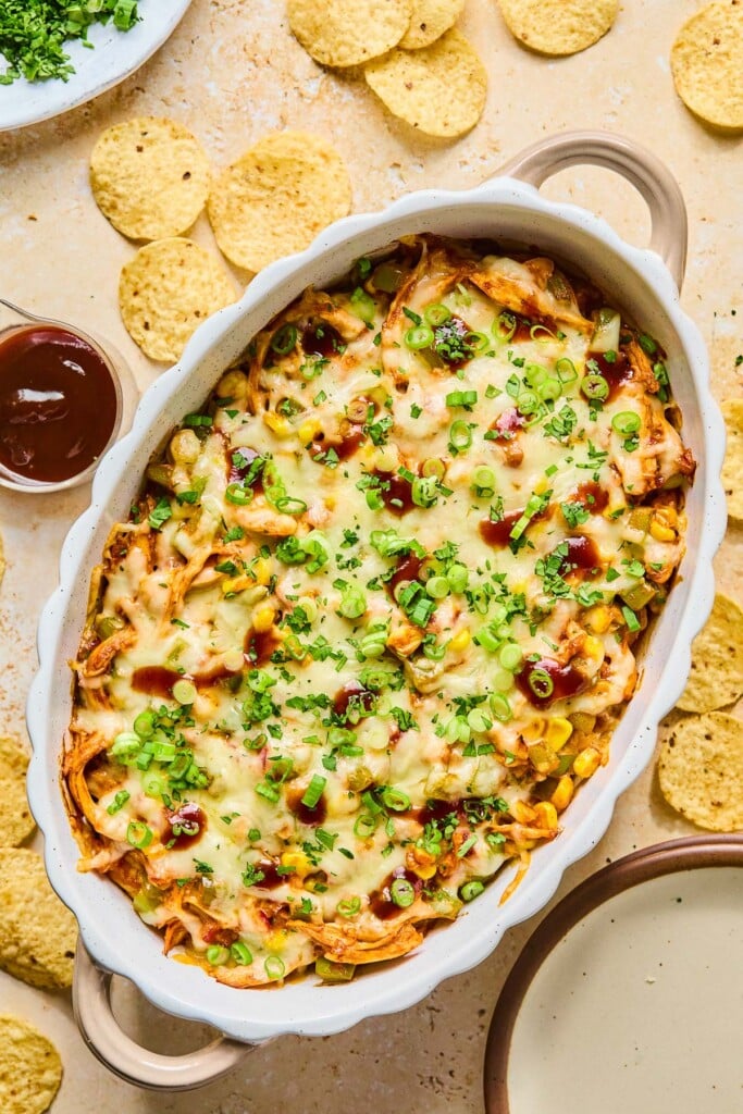 Au gratin dish filled with BBQ chicken dip topped with melted cheese and garnished with sliced green onions, cilantro, and BBQ sauce. Around the dish are some tortilla chips, an empty plate, a cup of bbq sauce, and a plate of cilantro.