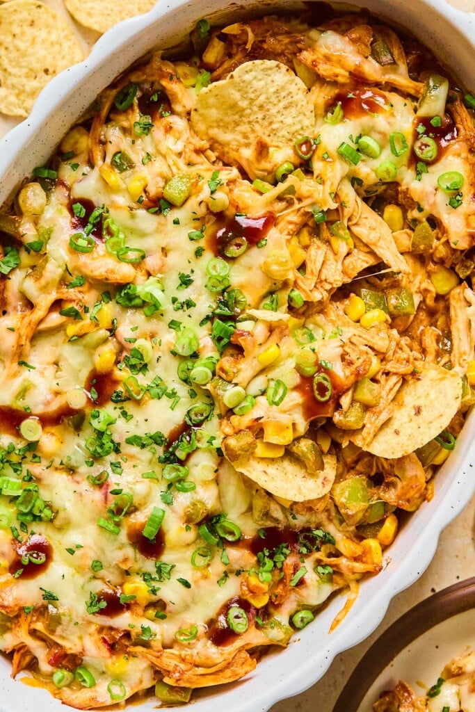 A close up of barbecue chicken dip topped with melted cheese and garnished with sliced green onions, cilantro, and BBQ sauce in a white baking dish. Some tortillas chips are in the dip as well.
