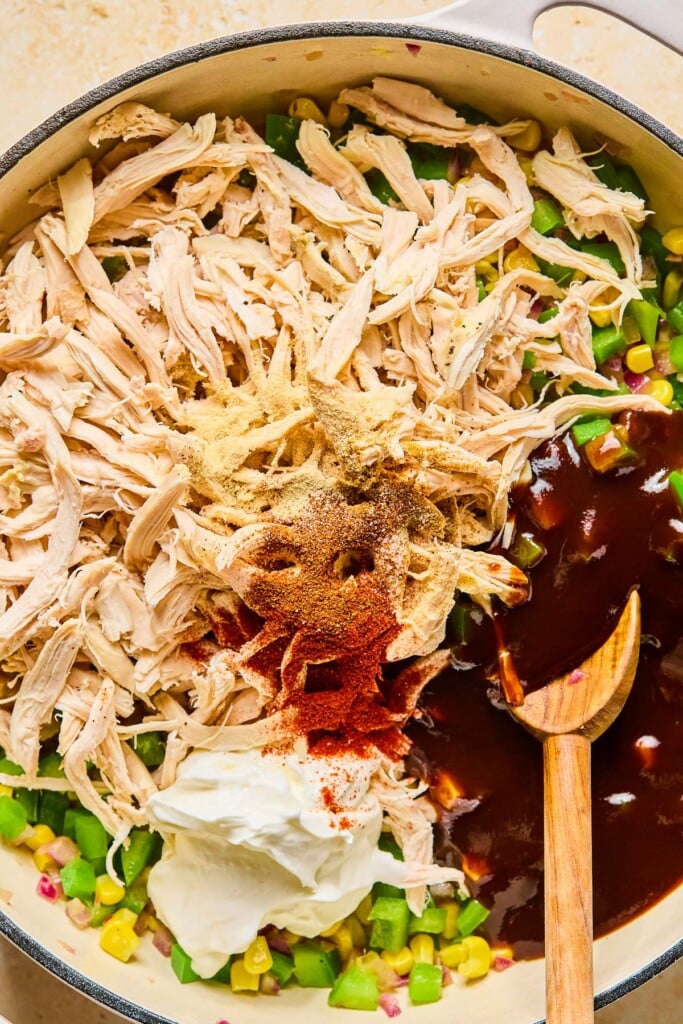 Large enameled pan filled with diced saut&eacute;ed veggies, shredded chicken, spices, greek yogurt, and BBQ sauce before it is mixed together. A wooden spoon is in the pan as well.