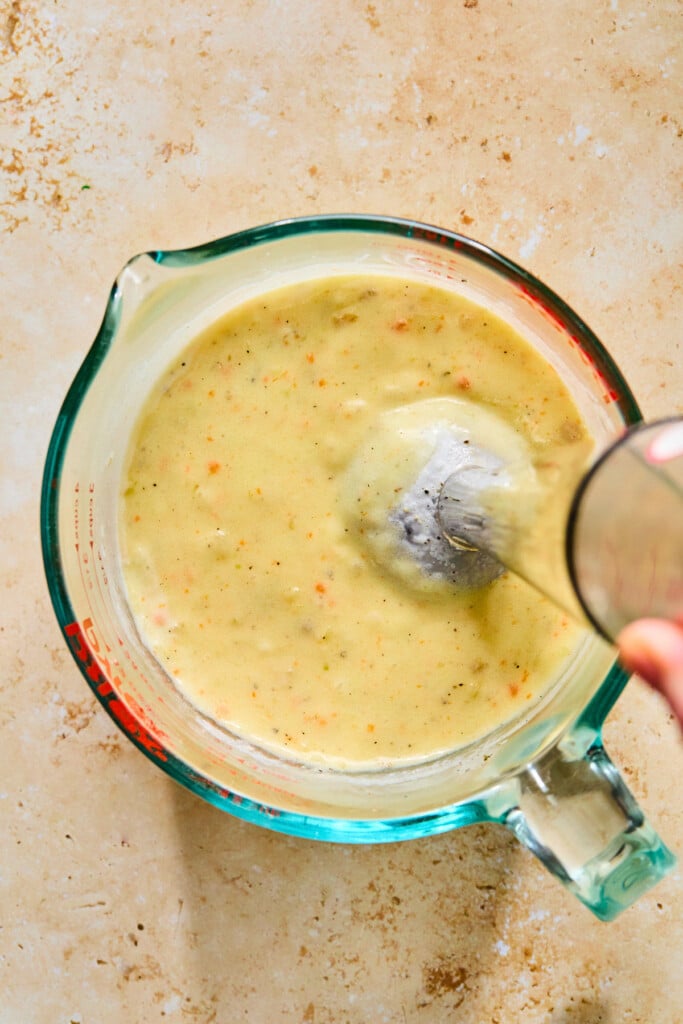 Measuring cup with a thick and creamy off-white broth made up of potatoes, veggies, and cream in it. An immersion blender is also in the measuring cup.