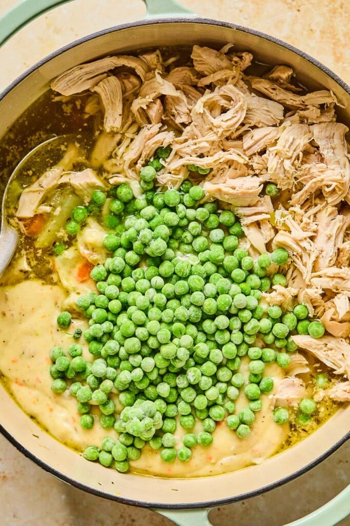 Large dutch oven filled with cooked potatoes and veggies in broth. Sitting on top on the broth is shredded chicken, a thick and creamy off-white broth, and frozen peas before it is mixed together. A large silver spoon is also in the pot.