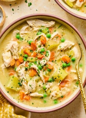 A bowl with healthy chicken pot pie soup with potatoes and veggies and garnished with fresh parsley and black pepper. A spoon is also in the bowl. Around the bowl is a mustard colored kitchen towel and another bowl of soup.