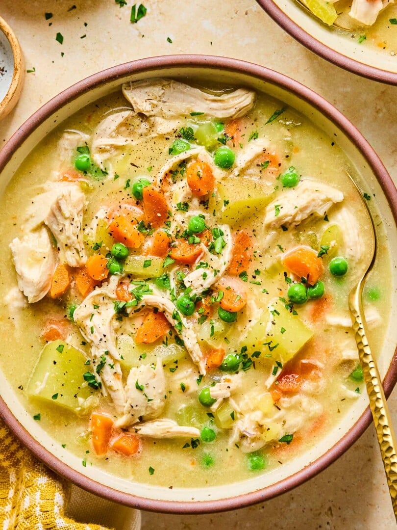 A bowl with healthy chicken pot pie soup with potatoes and veggies and garnished with fresh parsley and black pepper. A spoon is also in the bowl. Around the bowl is a mustard colored kitchen towel and another bowl of soup.