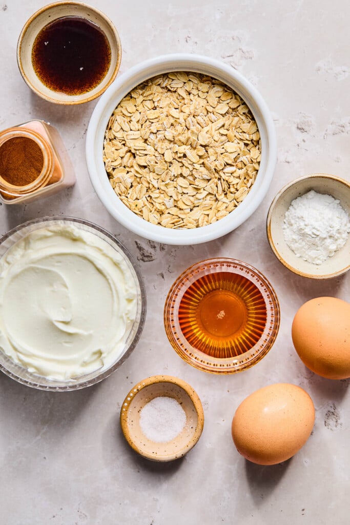 A bowl of rolled oats, a small bowl of baking powder, two large brown eggs, a small bowl of salt, a small bowl of honey, a bowl of greek yogurt, a spice jar of cinnamon, and a small bowl of vanilla bean paste on a creamy marble counter.
