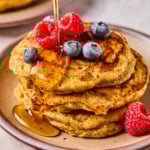 A stack of greek yogurt pancakes topped with raspberries and blueberries on a plate. Maple syrup is being poured on them. Around the plate are more berries.