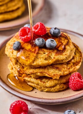 A stack of greek yogurt pancakes topped with raspberries and blueberries on a plate. Maple syrup is being poured on them. Around the plate are more berries.