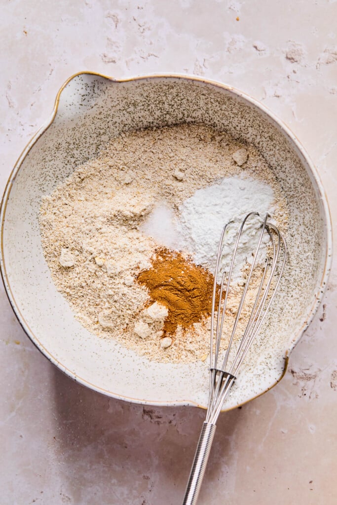 An overhead shot of a bowl with oat flour, cinnamon, baking powder, and salt before it is mixed together. A whisk is also in it.