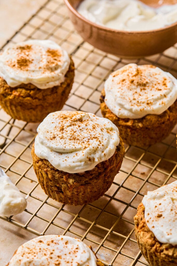 Carrot cake muffins topped with greek yogurt frosting and cinnamon on a wire cooling rack. There is also a bowl of icing on the rack.