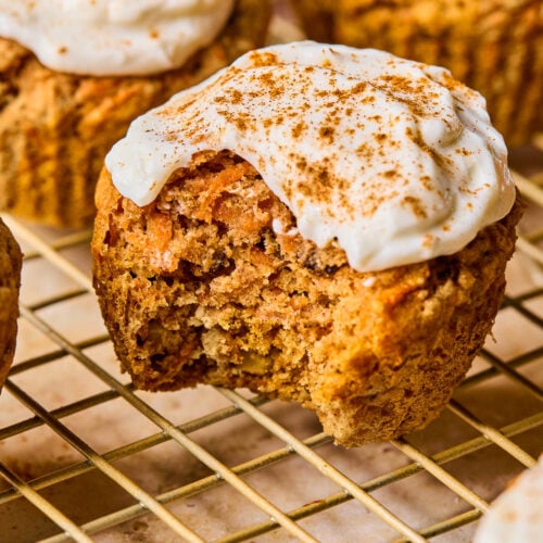 Healthy carrot cake muffins with greek yogurt frosting on a wire cooling rack. The one in front has a bite taken out of it.